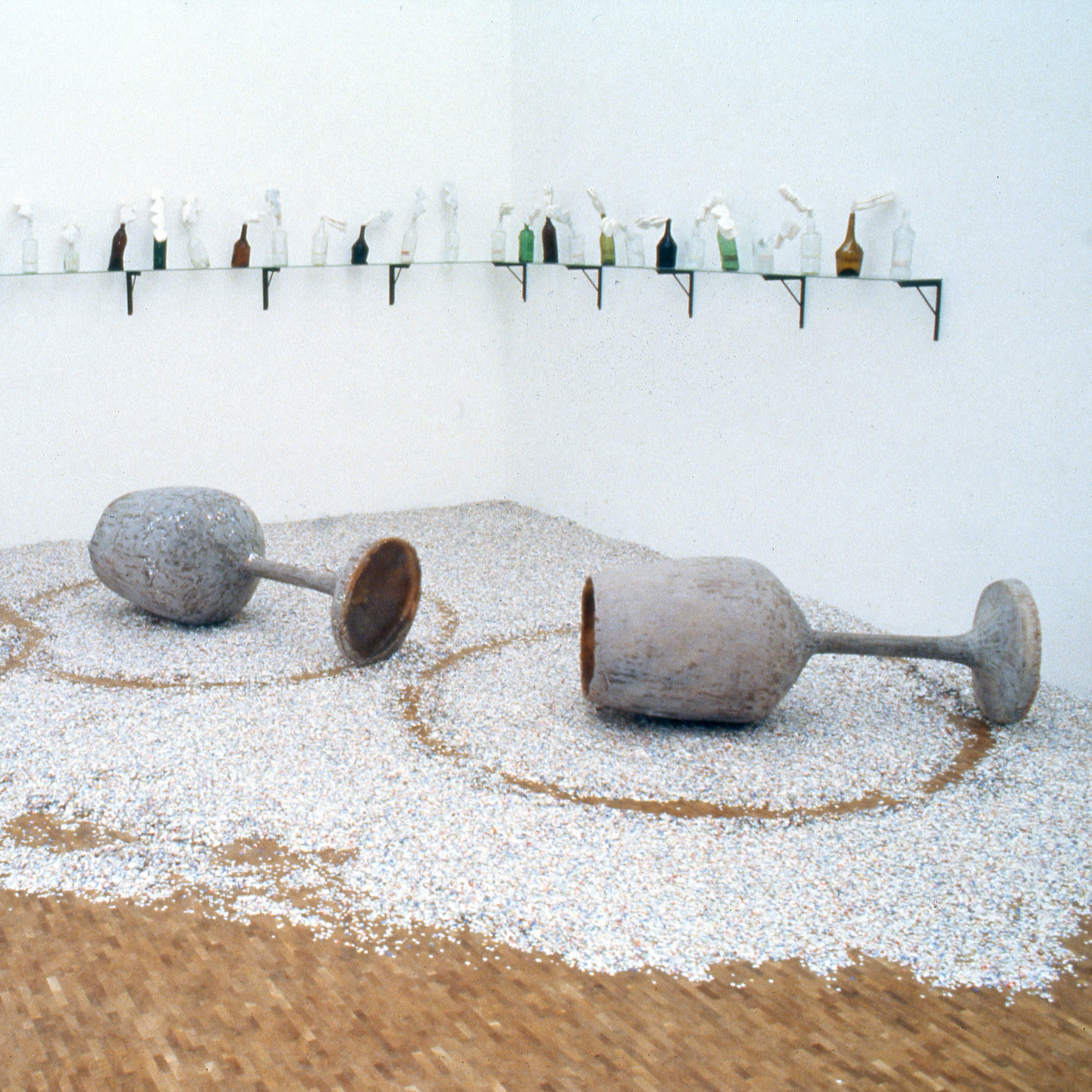 Photographie d'installation au centre d'une salle d'exposition. Représentant des verres à vin géants et des bouteilles sur des étagères aux murs.