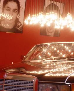 Salle d'exposition rouge avec portraits photographiques aux murs et une voiture sous un luminaire au centre.
